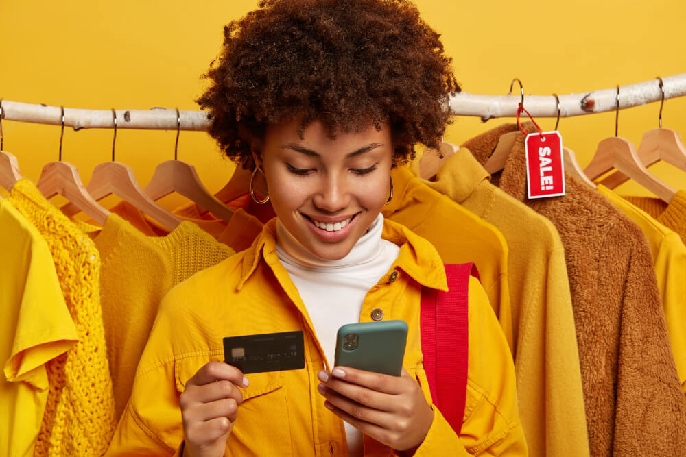 dark-skinned-woman-uses-modern-mobile-phone-credit-card-does-online-shopping-makes-order-via-internet-inserts-bank-account-information-stands-against-clothing-racks