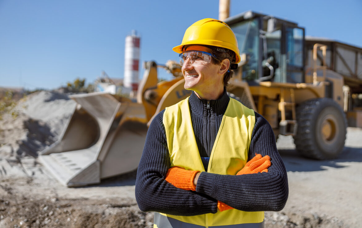 Joyful male engineer wearing safety helmet, glasses and vest while keeping arms crossed and smiling