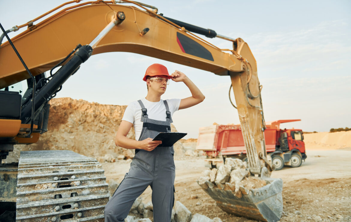 In orange colored hard hat. Worker in professional uniform is on the borrow pit at daytime.